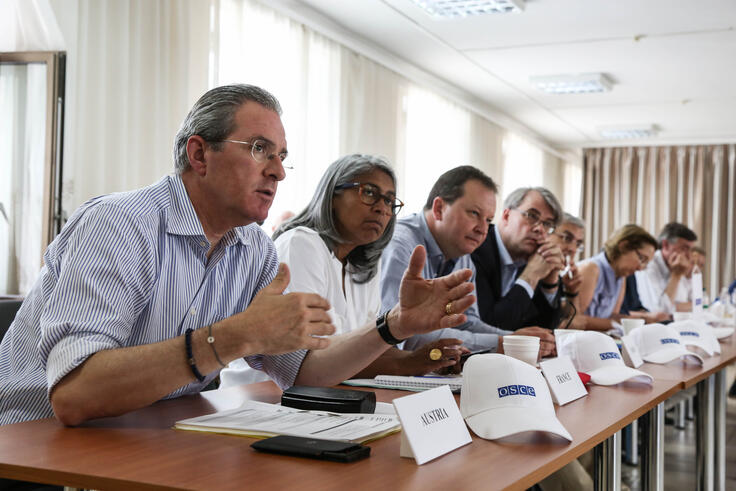 Several individuals, including men and women, sit at a conference table engaged in a discussion. The man in the foreground is speaking, gesturing with his hands, while others listen attentively. Small nameplates and white hats with the OSCE logo are visible on the table.
