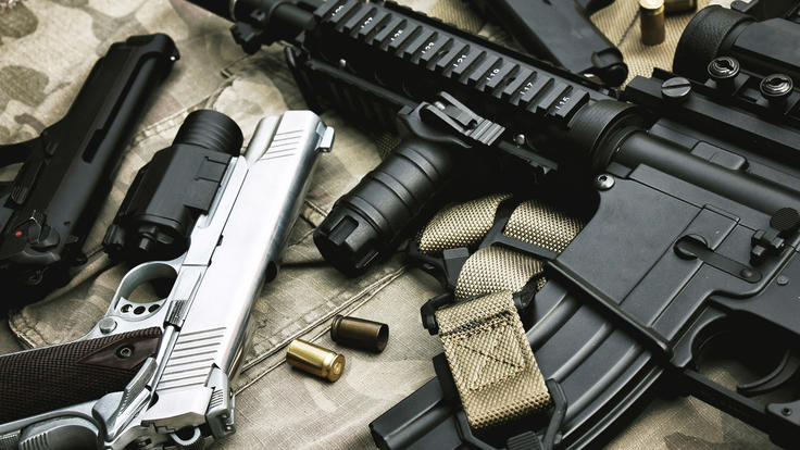 Close-up image of various firearms and ammunition placed on a camouflage-patterned surface.