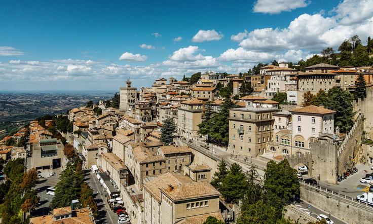 Hilltop town with tan buildings under a blue sky, surrounded by trees, and expansive landscape views.