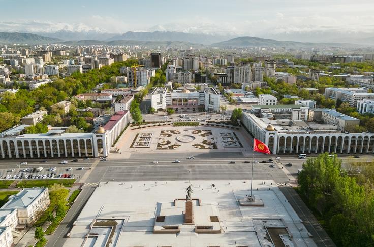Urban landscape with large square, surrounded by greenery and buildings, mountains in the distance, red flag in foreground.