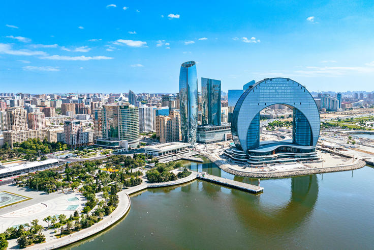 Aerial view of a modern cityscape with a circular skyscraper by a river under a clear blue sky.