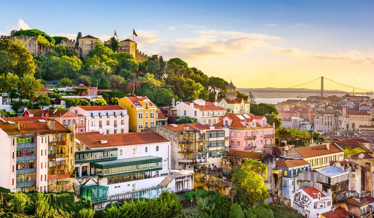 Colorful hillside homes in Lisbon with trees and a castle atop, a bridge in the background at sunset.