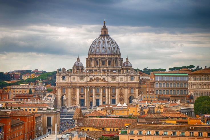 St. Peter's Basilica under a cloudy sky, surrounded by city buildings.