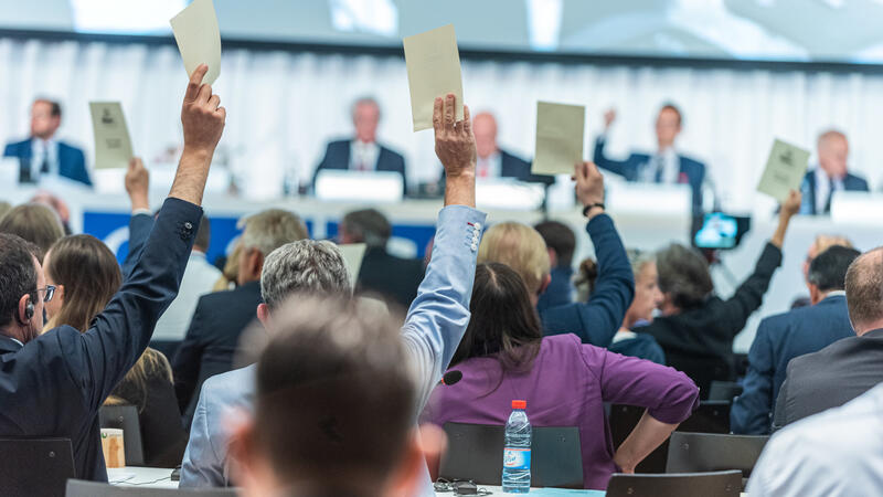 A group of people at a formal meeting raising voting cards, with a panel of speakers in the background.